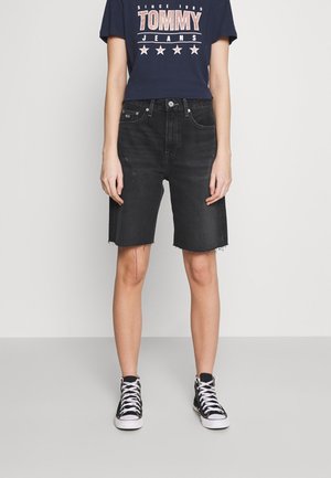 Person iført sorte, højtaljede denimshorts med rå kanter, navyblå grafisk t-shirt og sorte høje sneakers med hvide snørebånd og såler.