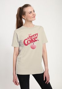 Světle béžové bavlněné tričko s grafickým potiskem "Užij si cherry Coke" v červené barvě, doplněné ikonou třešně se pruhovaným detailem.
