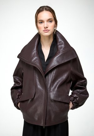 Jeune femme les mains dans les poches, portant une grande veste en cuir marron foncé à large col par-dessus un haut noir.