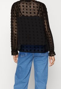 Blusa nera semi-trasparente con motivo a pois in rilievo; maniche lunghe a sbuffo; parte posteriore leggermente più lunga; design a strati; abbinata a jeans in denim azzurro.