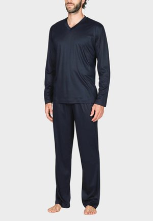 Uomo in piedi a piedi nudi che indossa una maglia blu navy scuro a manica lunga con scollo a V e pantaloni larghi abbinati, su uno sfondo chiaro e semplice.