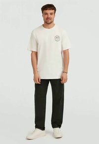 Man standing wearing white graphic T-shirt, black pants, white sneakers, and a bracelet against plain light background.