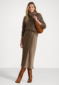 Hnedý pletený sveter s golierom, béžová midi sukňa a hnedé čižmy na podpätku po členky. Doplnky zahŕňajú béžovú kabelku na rameno s zakriveným tvarom.