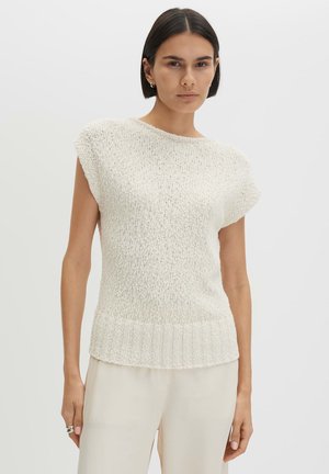 Frau mit kurzen dunklen Haaren, die ein strukturiertes weißes ärmelloses Stricktop und cremefarbene Hosen vor einem schlichten hellen Hintergrund trägt.