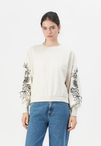Donna con capelli scuri in una felpa color crema con ricami floreali astratti neri sulle maniche, abbinata a jeans blu, in piedi su uno sfondo bianco.