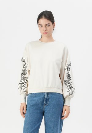 Kvinne med mørkt hår i en kremfarget genser med svart abstrakt blomsterbroderi på ermene, kombinert med blå jeans, stående mot en hvit bakgrunn.