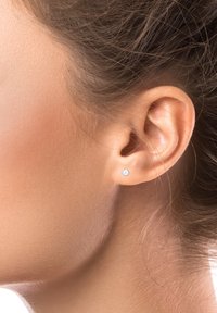 Arete de plata con una pequeña gema redonda. Diseño minimalista, contra un fondo neutro, que resalta la textura de la piel y la forma de la oreja.