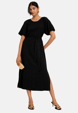 Schwarzes Maxikleid mit Puffärmeln, rundem Ausschnitt und gebundenem Taillengürtel. Mit einem seitlichen Schlitz und kombiniert mit schwarzen Sandalen und einer goldenen Clutch.