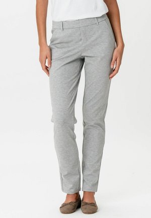 Femeie purtând pantaloni gri deschis, croi slim, asortați cu balerini maro, în picioare pe un fundal simplu alb.