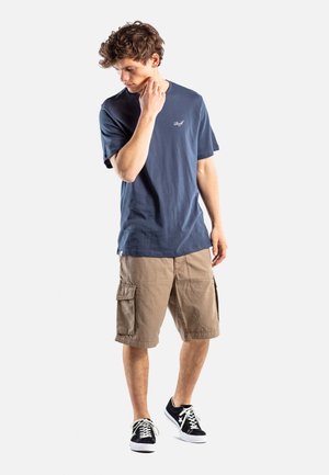 Jonge man die een donkerblauw t-shirt, beige cargoshorts en zwarte sneakers draagt, staand terwijl hij zijn nek aanraakt en naar beneden kijkt.