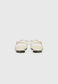Flate sandaler med en beige syntetisk overdel som har tynne stropper og en liten spenne. Sålen er svart og glatt, noe som gir et minimalistisk design.