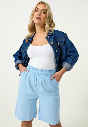 Kvinde iført en blå denimjakke over en hvid tanktop og lyseblå shorts med elastisk talje, stående med hænderne i lommerne.