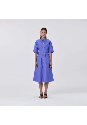 Femme debout sur un fond uni portant une robe bleue boutonnée jusqu'aux genoux, manches courtes, ceinture large et sandales noires.