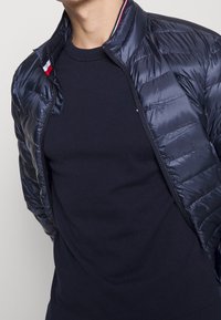 Marineblaue gesteppte Jacke mit glatter Textur und horizontalen Steppnähten, ausgestattet mit einem hohen Kragen und einem bunten Akzent im Innenbereich des Kragens.