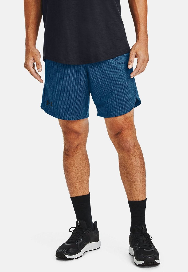 Homme portant un short de sport bleu en mesh, des chaussettes noires mi-mollet, des chaussures de course noires et un t-shirt noir, debout sur un fond blanc.
