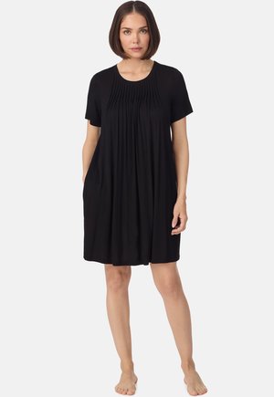 Frau mit kurzen braunen Haaren, die ein lockeres schwarzes, knielanges Kleid mit kurzen Ärmeln und Falten auf der Brust trägt, steht barfuß vor weißem Hintergrund.