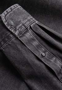 Zwarte denim stof met een gestructureerd oppervlak, met een geplooid ontwerp en twee zwarte drukknopen bij de kraag.