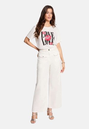 Femme debout, main sur la hanche, portant un pantalon blanc à jambes larges, un t-shirt blanc à motif avec le mot "LOVE" et des lèvres rouges, et des talons argentés ouverts à l'avant.