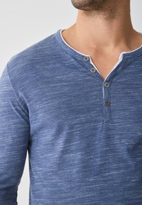 Koszula z długim rękawem w stylu henley, uszyta z teksturowanej niebieskiej tkaniny, z białym kołnierzykiem. Ma trzy ciemne guziki przy dekolcie.