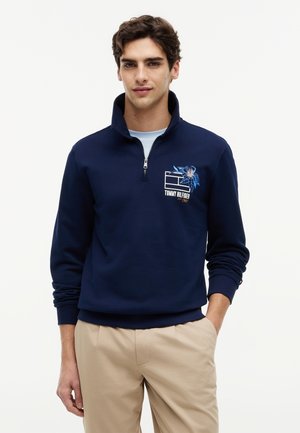 Fiatal férfi tengerészkék negyedcipzáras pulóverben, virágos logóval és bézs nadrágban, egyik kezével zsebre dugva, fehér háttér előtt áll.