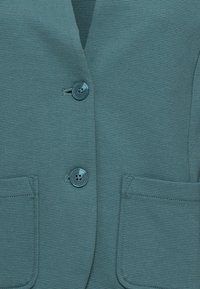 Blazer azul verdoso con una textura acanalada, dos botones y dos bolsillos frontales. Los detalles notables incluyen botones a juego y costuras limpias.