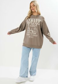 Sudadera grande marrón con texto gráfico "Coffee Club", corte relajado, puños y dobladillo acanalados, combinada con jeans rectos de tono azul claro y zapatillas blancas.