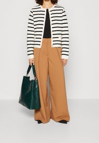 Cardigan a righe con linee orizzontali bianche e nere, abbinato a pantaloni beige a gamba larga. Tiene in mano una borsa in pelle scura verde con texture.