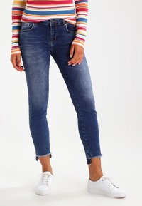 Ljusblå denimjeans med slim fit, fransade sömmar och subtila blekningar, kombinerade med vita sneakers och en långärmad topp med flerfärgade ränder.