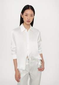 Chemise en satin blanc à boutonnières avec un col italien et des manches retroussées, associée à un pantalon en argent brillant, créant un look moderne et élégant.