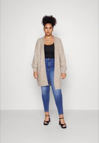 Cardigã de malha bege com mangas longas, combinado com uma blusa preta e jeans skinny azuis. O modelo usa sandálias de salto pretas.