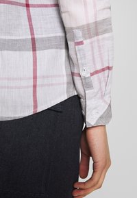 Manche d'une chemise à carreaux blanc, gris et rouge boutonnée au poignet, reposant à côté d'un pantalon en denim foncé et d'une main détendue.