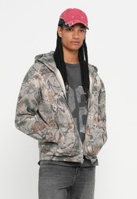 Jaunietis ar gariem aizsietiem matiem, uzsliets sarkans cepure, puscamo veida kapuci ar kamuflāžas rakstu, tumšu grafikas t-kreklu un pelēkām biksēm, stāv ar rokām kabatās.