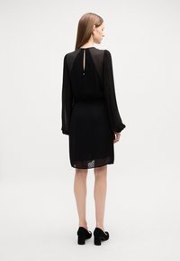 Rochie neagră cu mâneci lungi translucide, textură cu buline și detaliu cu decupaj la spate. Asortată cu pantofi cu toc bloc din suede negru.