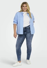 Camisa de botones a rayas azul claro y blanca sobre una camiseta blanca, combinada con unos jeans ajustados azul oscuro y zapatillas blancas, en una pose de pie.