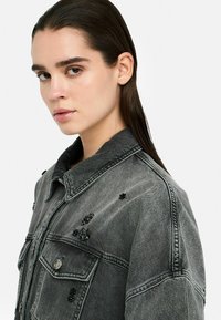 Graue Jeansjacke mit Kragen, verziert mit schwarzen Perlen und zwei Fronttaschen. Der Stoff hat eine abgenutzte Textur.