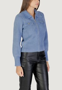 Cardigan in maglia fuzzy di colore blu chiaro con chiusura a zip, colletto a punta e due tasche sul petto. Abbinato a pantaloni in pelle nera.