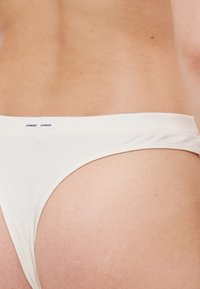 Bijela pamučna tanga s mekim pojasom i malim oznakom branda. Tkanina ima glatku teksturu i minimalistički dizajn.