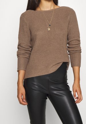 Vrouw die een bruin geribbeld gebreide trui met lange mouwen draagt en zwarte nepleren broek, gecompleteerd met twee gouden kettingen.