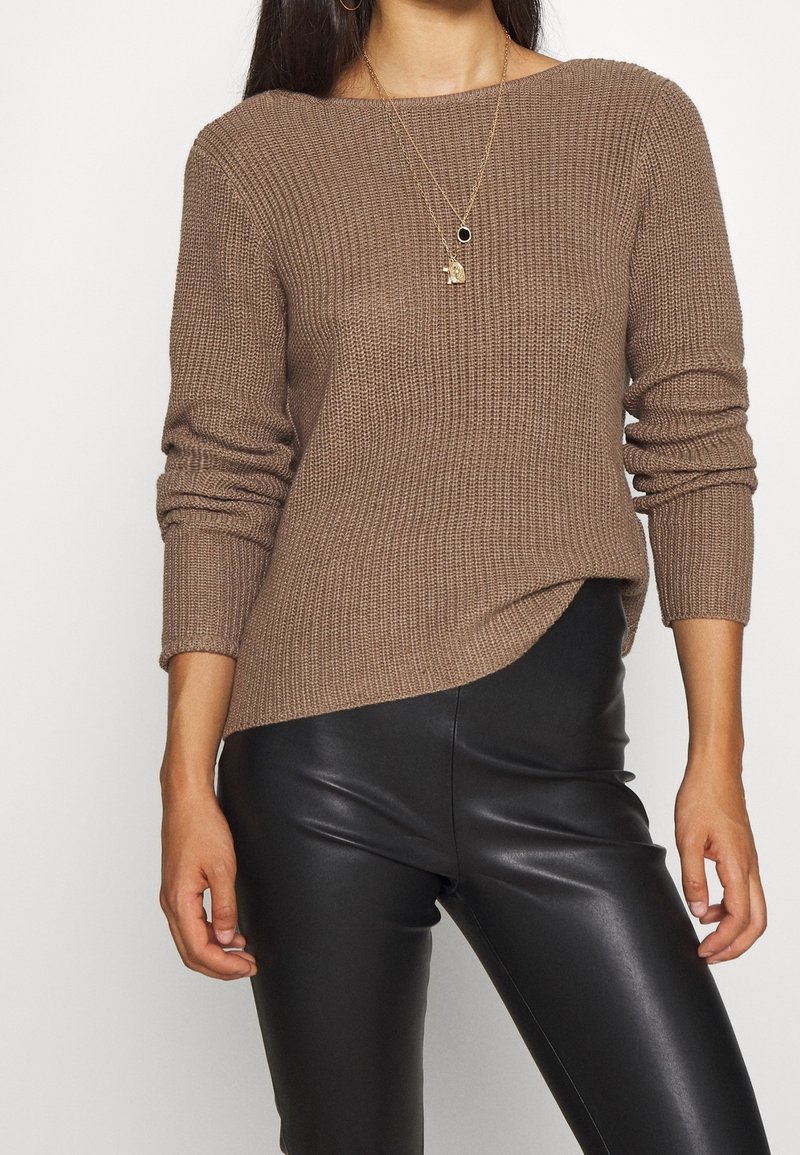 Femme portant un pull en maille côtelée marron à manches longues et un pantalon en cuir synthétique noir, accessoirisée de deux colliers en or.