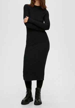 Kvinde iført en lang sort ribstrikket kjole, sorte leggings og sorte snørebånds-støvler, stående med armene krydsede.