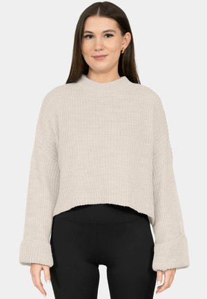 Junge Frau mit langen braunen Haaren, die einen beigen grob gestrickten Pullover mit übergroßen Ärmeln und schwarze Leggings mit hoher Taille trägt.