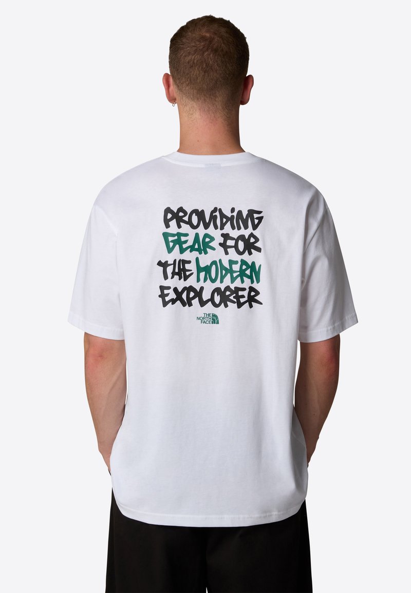 Uomo con i capelli corti e un orecchino che indossa una maglietta bianca con testo verde e nero "Fornire attrezzature per l'esploratore moderno" e il logo The North Face.