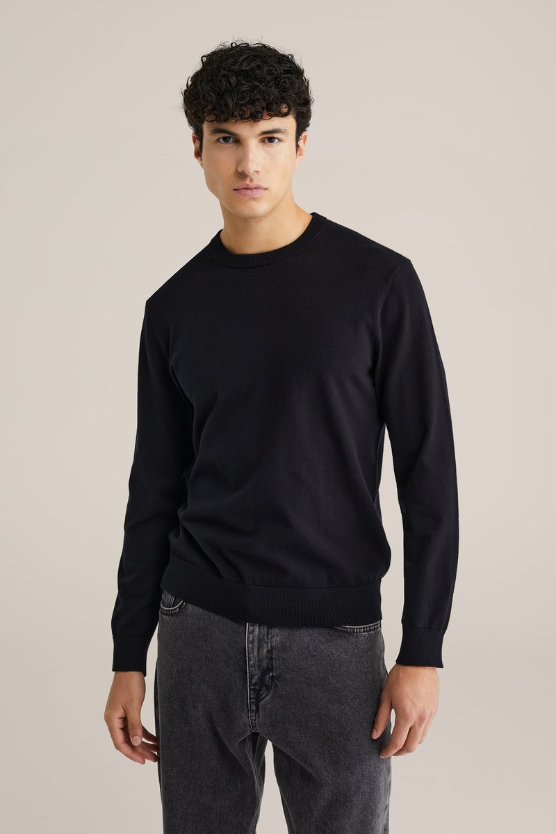 Melna kokvilnas džemperis ar garām piedurknēm, ribotām aprocēm un apakšmalu; kombinēts ar pelēkām džinsām. Vienkāršs, pieguļošs dizains bez rakstiem.