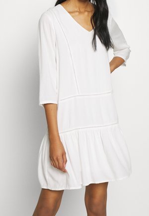 Femme portant une robe ample blanche atteignant les genoux, avec des manches trois-quarts, un col en V et une jupe froncée, sur un fond uni.