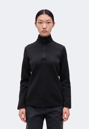 Une femme porte un pull noir texturé à col montant zippé et un pantalon foncé sur un fond clair et uni.