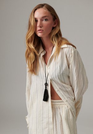 Jeune femme aux longs cheveux blonds porte une chemise ample rayée crème et un pantalon assorti, accessoirisés d’un collier noir à pompons.