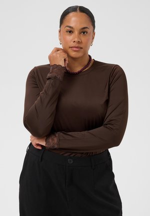 Femme aux cheveux foncés en chignon bas porte un haut marron à manches longues avec des poignets en dentelle et un pantalon noir, accessoirisé d’un collier de perles violet.