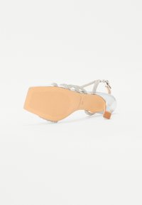 Silberne High-Heel-Sandale mit einem strukturierten Riemen, quadratischem Absatz und glatter Sohle. Die Unterseite verfügt über erhabene Linien für besseren Halt.