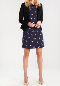 Laivastonsininen napillinen mekko, jossa on flamingokuvio, yhdistetty istuvaan mustaan bleiseriin ja metallinhohtoisiin korkokenkiin, ja siinä on sileä kangaspinta.