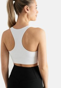Valkoinen urheilullinen crop-top, jossa on takana urheilurintaliivityyppinen muotoilu, tasaisia saumoja ja sileä rakenne. Yhdistetty korkeavyötäröisten mustien leggingsien kanssa.